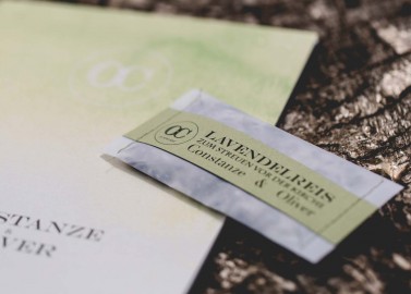 Hochzeitswahn - Eine Nahaufnahme eines kleinen Päckchens Lavendeltee mit der Aufschrift „Lavendel Tees“, das auf einer strukturierten Baumrinde liegt, neben einem hellgrünen Papier mit einem verblassten runden Logo.