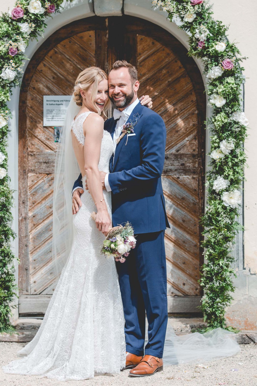 Hochzeitswahn - Eine Braut und ein Bräutigam lächeln freudig vor einer kunstvoll verzierten Holztür, die mit Blumendekorationen geschmückt ist. Die Braut trägt ein langes weißes Spitzenkleid und einen Schleier und hält einen Blumenstrauß, während der Bräutigam einen blauen Anzug mit einer Ansteckblume trägt.
