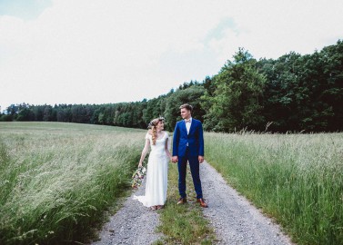 Hochzeitswahn - Braut und Bräutigam gehen Hand in Hand einen Feldweg entlang einer üppigen Wiese mit Bäumen und klarem Himmel im Hintergrund. Die Braut trägt eine weiße Blumenkrone und einen Blumenstrauß; der Bräutigam trägt einen schicken blauen Anzug.