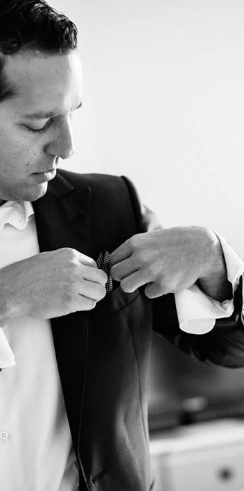 Hochzeitswahn - Ein Graustufenbild eines Mannes, der einen dunklen Anzug und ein weißes Hemd trägt und in einem Raum mit minimalistischem Dekor steht, während er seine Manschettenknöpfe zurechtrückt.