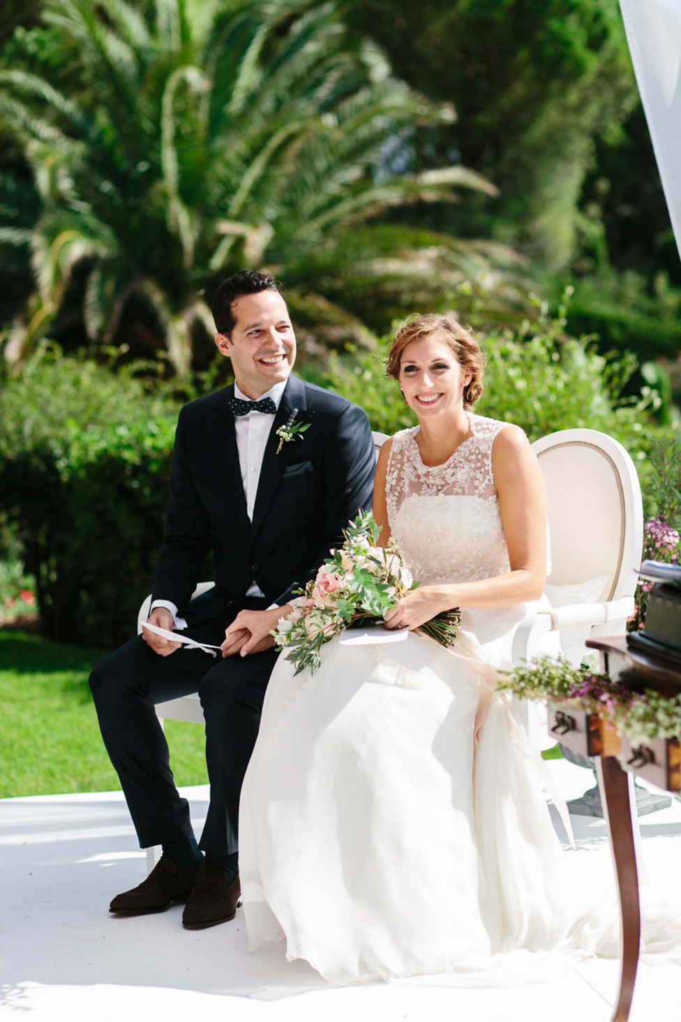 Hochzeitswahn - Braut und Bräutigam sitzen während ihrer Hochzeitszeremonie nebeneinander im Freien und lächeln. Die Braut hält einen Strauß zartrosa Blumen und beide sind elegant in Abendgarderobe gekleidet. Im Hintergrund ist üppiges Grün zu sehen.