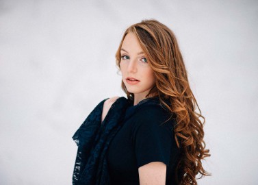 Hochzeitswahn - Eine junge Frau mit langen, lockigen roten Haaren und heller Haut blickt vor einem sanften weißen Hintergrund über ihre Schulter. Sie trägt eine dunkle Bluse und hat einen nachdenklichen Gesichtsausdruck.