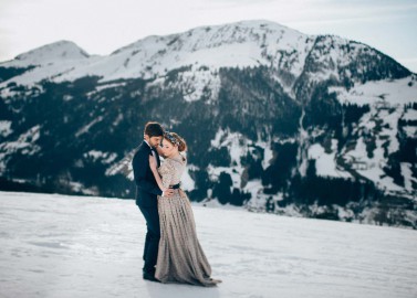 Hochzeitswahn - Ein Paar in Abendgarderobe umarmt sich in einer verschneiten Berglandschaft mit markanten schneebedeckten Gipfeln im Hintergrund und vermittelt so eine romantische Winterszene.