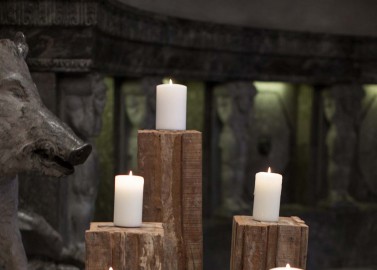 Hochzeitswahn - Kerzen auf unterschiedlich hohen rustikalen Holzkerzenhaltern in einem schwach beleuchteten Raum mit klassischen Skulpturen im Hintergrund schaffen eine ruhige und besinnliche Atmosphäre.