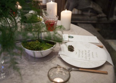 Hochzeitswahn - Ein elegant gedeckter Tisch mit einer Menükarte, edlem Besteck, einem Cocktailglas und mehreren brennenden Kerzen inmitten einer grünen Kulisse, die eine warme, intime Atmosphäre beim Essen schafft.