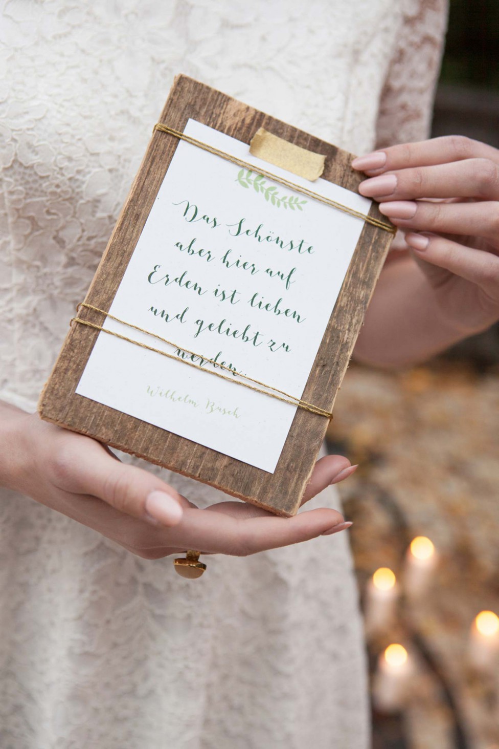 Hochzeitswahn - Eine Braut in einem Spitzenkleid hält ein holzgerahmtes Hochzeitsschild mit einem Zitat von Wilhelm Busch in Schrift, akzentuiert durch einen grünen Zweig.