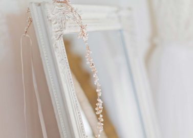 Hochzeitswahn - Ein zartes Brauthaarteil aus Perlen und Kristallen hängt über der Ecke eines verzierten weißen Spiegelrahmens, vor einem weichgezeichneten cremefarbenen Hintergrund, und vermittelt ein Gefühl der Eleganz des Hochzeitstages.