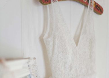Hochzeitswahn - Ein weißes Brautkleid mit Spitzenmieder und Tüllrock hängt an einer Tür und wird teilweise in einem an die Wand gelehnten, reich verzierten Vintage-Spiegel reflektiert. Der Fokus ist weich und die Beleuchtung sanft, was eine heitere Atmosphäre schafft.