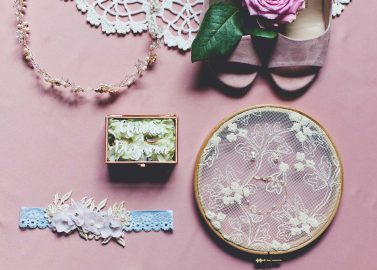 Hochzeitswahn - Eine flache Anordnung von Brautaccessoires auf einem rosa Hintergrund: eine Tiara, ein Spitzenstrumpfband, ein Blumenschmuck, eine Halskette und ein Stickrahmen mit Spitzendetails, neben einer Schachtel mit der Aufschrift „Always & Forever“.