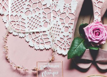 Hochzeitswahn - Ein Flatlay-Bild mit Brautaccessoires auf rosa Hintergrund. Zu den Artikeln gehören eine zarte Halskette, eine offene Ringschachtel mit der Aufschrift „Brautjungfer“ und Blütenknospen darin, eine rosa Rose, ein Spitzenstrumpfband und ein bestickter Reif.