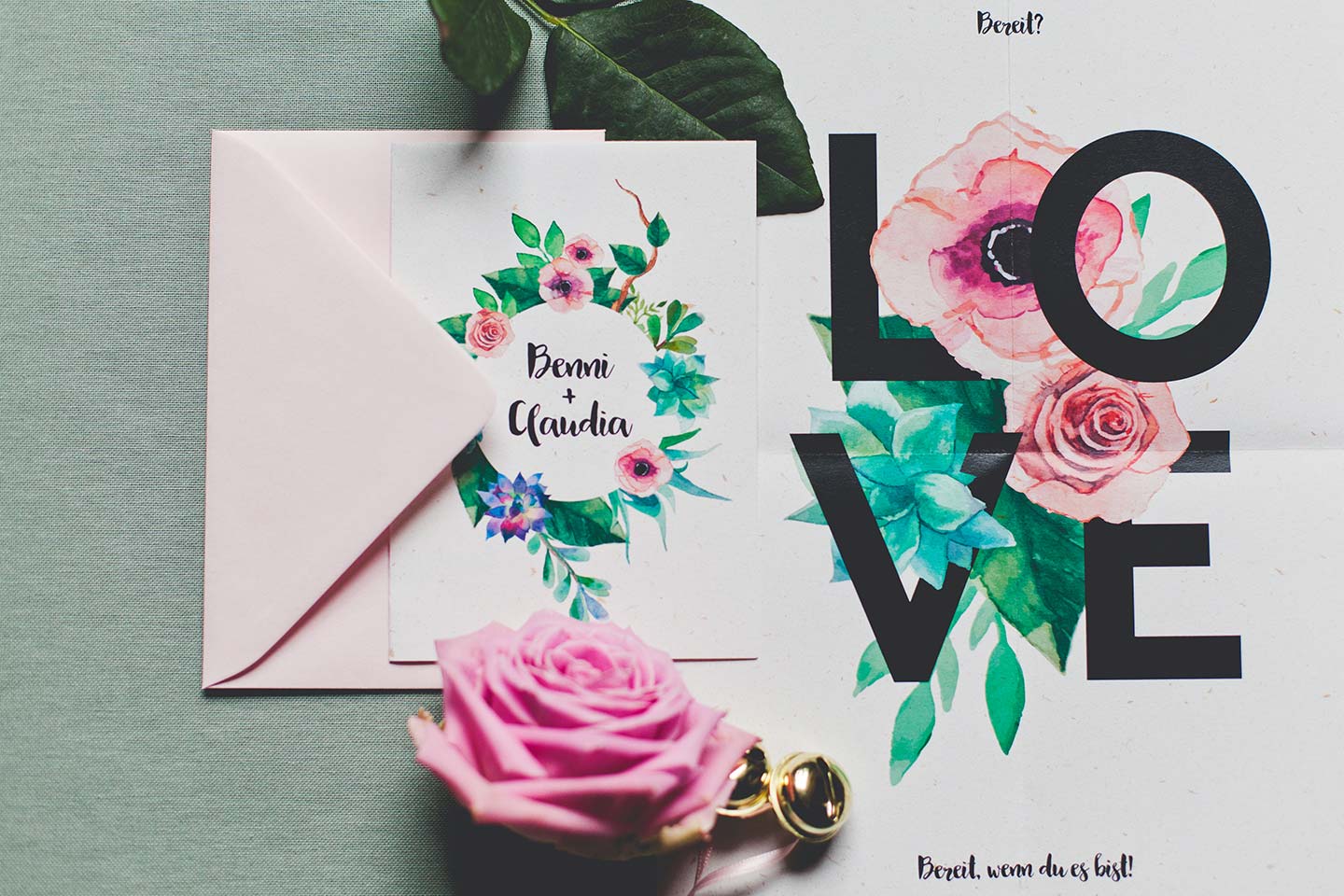 Hochzeitswahn - Eine Karte mit Blumenmotiv, auf der das Wort „Liebe“ in fetten schwarzen Buchstaben auf bunten Blumen neben einer blühenden rosa Rose und einem goldenen Ring liegt. Eine handschriftliche Notiz auf der Karte lautet „Beni & Claudia“.