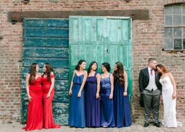 Hochzeitswahn - Eine Hochzeitsgruppe in farbenfroher Abendkleidung steht neben einem rustikalen Backsteingebäude mit einer grünen Holztür. Zwei Personen auf der rechten Seite küssen sich, während andere miteinander plaudern und lachen. Wasserburg-Geretzhoven