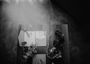 Hochzeitswahn - Schwarzweißfoto einer Hochzeitseinladung, einer Antwortkarte und einer Blumenansteckblume auf einer Holzoberfläche, beleuchtet von weichem, diffusem Licht.