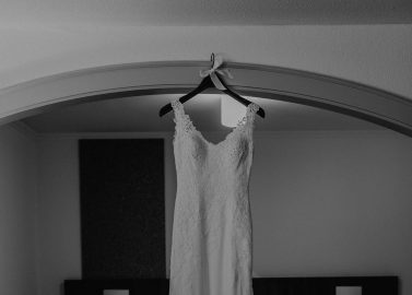 Hochzeitswahn - Ein Schwarzweißfoto eines Spitzenbrautkleides, das in einem Raum mit einem Bett und zwei Kissen im Hintergrund hängt. Das Kleid wird in einem gut beleuchteten Raum unter einer gewölbten Decke ausgestellt.