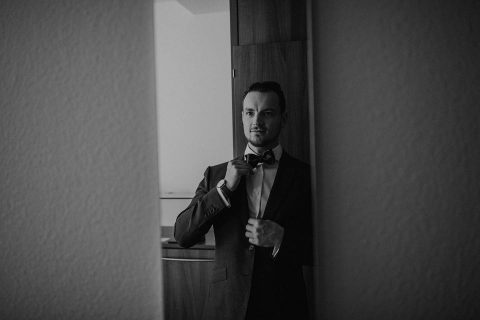 Hochzeitswahn - Ein Schwarzweißbild eines Mannes im dunklen Anzug, der neben einer offenen Tür steht und seine Krawatte zurechtrückt. Er wirkt konzentriert, im Spiegel hinter ihm ist ein leichtes Spiegelbild zu erkennen.
