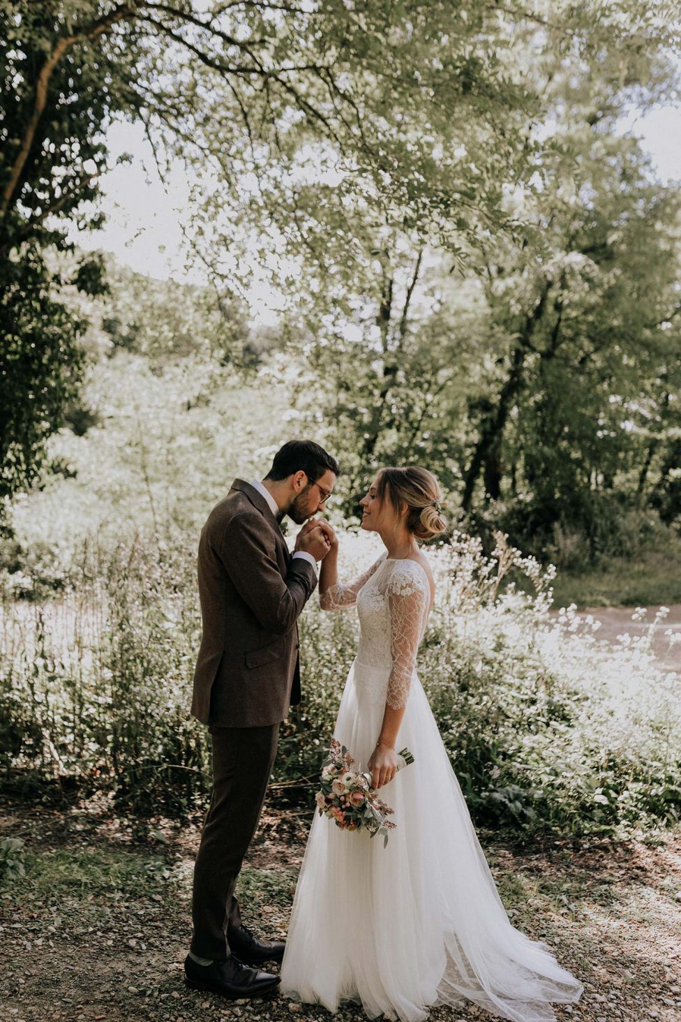 Hochzeitswahn - Ein zärtlicher Moment in einer üppigen Waldkulisse, als der Bräutigam seiner Braut, die einen Blumenstrauß hält, die Hand küsst. Beide sind für ihren Hochzeitstag elegant gekleidet.