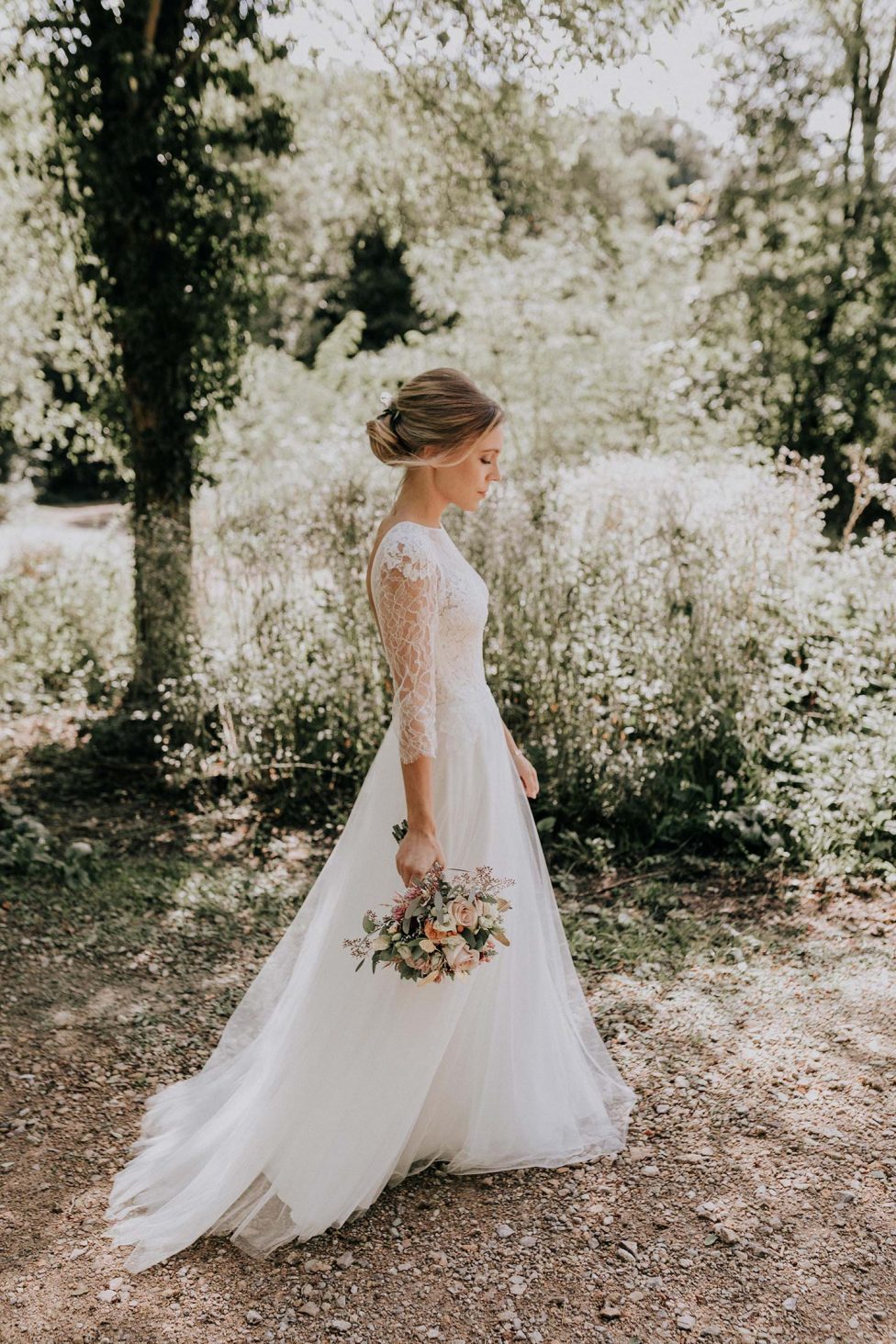 Hochzeitswahn - Eine Braut in einem eleganten Brautkleid mit Spitzenärmeln hält einen Blumenstrauß in der Hand, ihre Haare sind hochgesteckt, während sie nachdenklich im Freien spazieren geht, während das gesprenkelte Sonnenlicht durch die Bäume fällt.