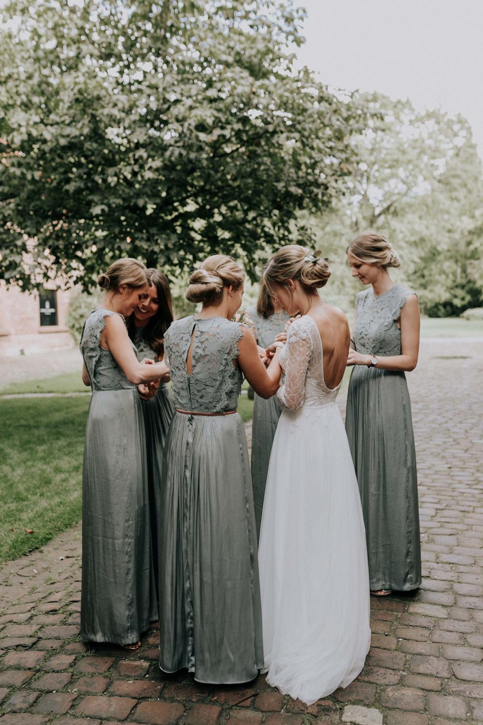 Hochzeitswahn - Vier elegant gekleidete Frauen – eine Braut in Weiß und drei Brautjungfern in Grau – teilen einen freudigen Moment miteinander, mit dem Rücken zur Kamera, und enthüllen komplizierte Kleiderdetails und eine warme, unterstützende Atmosphäre.