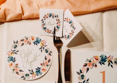 Hochzeitswahn - Ein elegant gedeckter Veranstaltungstisch mit dekorativen Menüs und Tischnummern im Blumenmuster sowie Silberbesteck auf einer strukturierten Tischdecke zeugt von einem sorgfältig geplanten Speiseerlebnis.