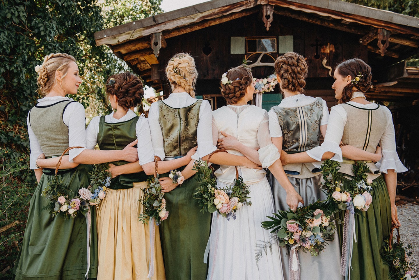 Hochzeitswahn - Eine Gruppe von Frauen in traditionellen Dirndlkleidern steht Arm in Arm mit Blumensträußen vor einer rustikalen Hütte und verkörpert Kameradschaft und kulturelles Erbe.