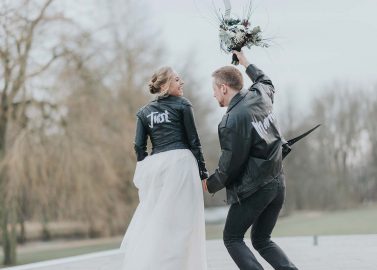 Hochzeitswahn - Eine verspielte Braut und ein verspielter Bräutigam, unkonventionell gekleidet mit Lederjacken über dem Hochzeitskleid, verbringen einen freudigen Moment im Freien, wobei der Bräutigam verspielt den Brautstrauß hochhebt, während sie sich anschauen und lächeln.