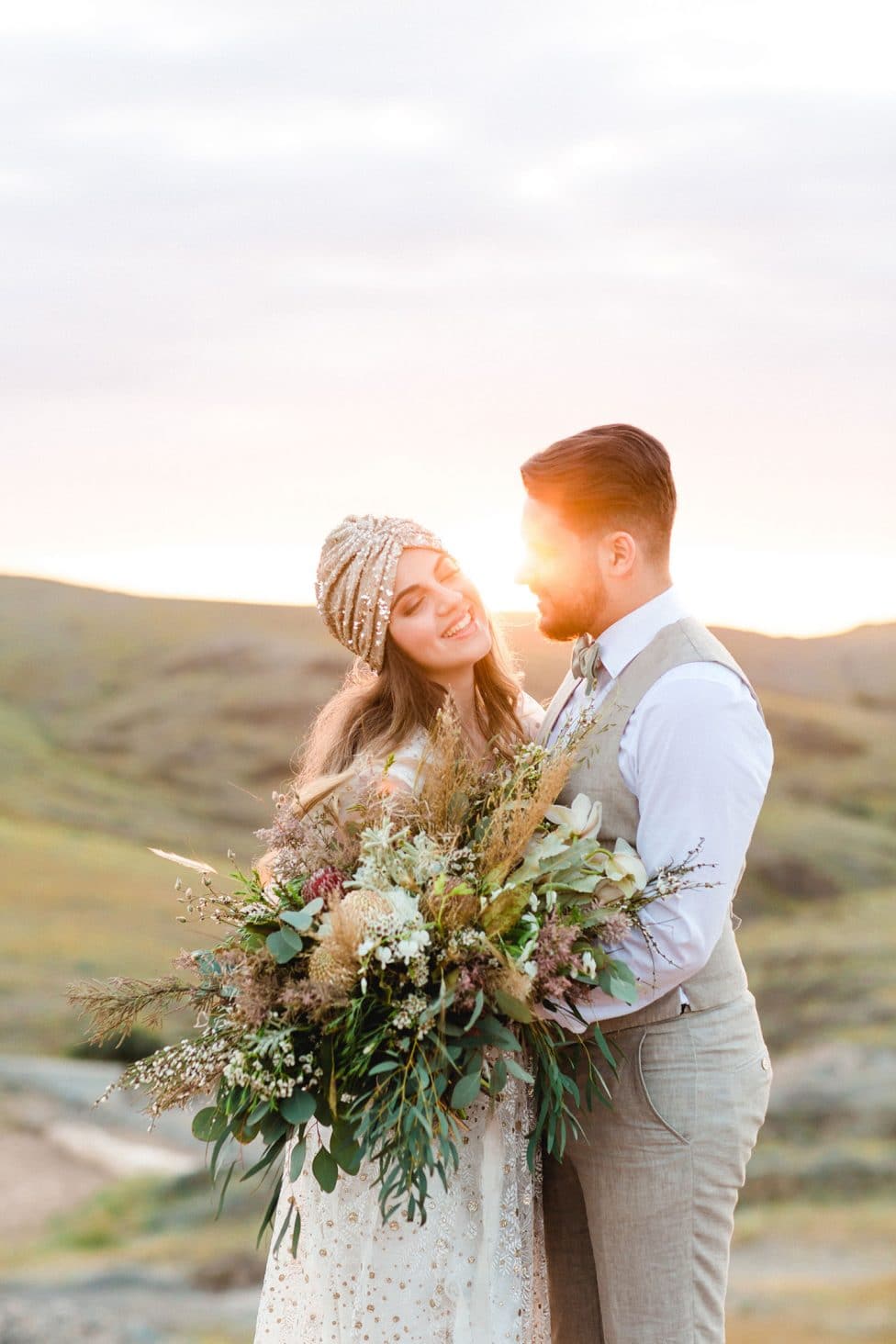 Hochzeitswahn - Ein strahlendes Paar in Hochzeitskleidung teilt einen zärtlichen Moment inmitten eines goldenen Sonnenuntergangs auf einem ruhigen Hügel, während die Braut einen großen, naturalistischen Blumenstrauß hält.