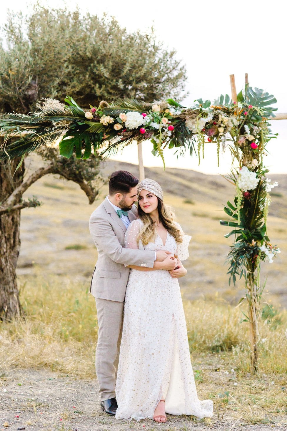 Hochzeitswahn - Ein Paar umarmt sich liebevoll unter einem floralen Hochzeitsbogen in einer ruhigen Umgebung im Freien und präsentiert seine Hochzeitskleidung im Bohème-Stil vor dem Hintergrund sanfter Wildnis.