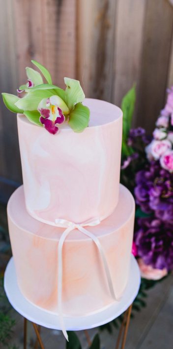 Hochzeitswahn - Eine zweistöckige, elegant gestaltete Torte mit rosafarbenem Zuckerguss und dezenten weißen Wirbeln, gekrönt von einer frischen grünen Orchidee, wird von einem Strauß leuchtend violetter Blumen getragen und verkörpert eine perfekte Mischung aus Raffinesse und botanischem Charme.