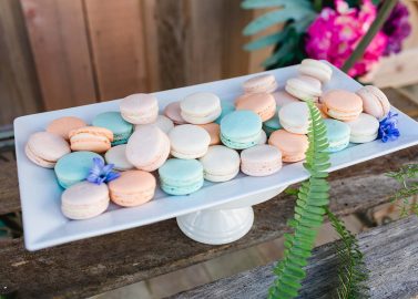 Hochzeitswahn - Eine Reihe pastellfarbener Macarons, geschmückt mit kleinen Blumen, elegant präsentiert auf einer weißen rechteckigen Platte, vor einem rustikalen Holzhintergrund mit lebendigen Blumenakzenten.