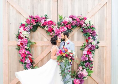 Hochzeitswahn - Ein Paar teilt einen Kuss, umrahmt von einem herzförmigen Blumenarrangement auf einer Holzkulisse, der Romantik bei ihrer Hochzeitsfeier verkörpert.