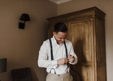 Hochzeitswahn - Ein Mann in weißem Hemd, Hosenträgern und heller Hose lächelt, während er in einem warm beleuchteten Raum mit einem Holzschrank und einer kleinen Lampe im Hintergrund seine Hosenträger zurechtrückt. Schloss-Langenburg