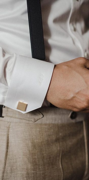 Hochzeitswahn - Ein Mann in formeller Kleidung richtet seine Manschettenknöpfe. Der Fokus liegt auf seinen Händen, während er ein weißes Oberhemd, graue Hosen und einen schwarzen Gürtel trägt. Schloss-Langenburg