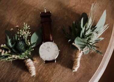 Hochzeitswahn - Eine Armbanduhr mit weißem Zifferblatt und braunem Lederarmband liegt neben zwei kleinen Sträußen aus Grünpflanzen und Disteln, die alle auf einer Holzoberfläche arrangiert sind. Schloss-Langenburg