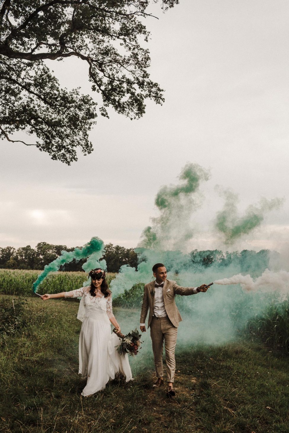 Hochzeitswahn - Braut und Bräutigam halten bunte Rauchbomben in den Händen und laufen durch eine Wiese, während Rauch in türkisfarbenen und grünen Farbtönen um sie herumwirbelt und eine verträumte Atmosphäre schafft. Schloss-Langenburg