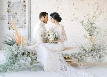 Hochzeitswahn - Ein frisch verheiratetes Paar erlebt einen intimen Moment, umgeben von einer ruhigen Umgebung aus weißen Blumen und rustikalem Backstein, die eine schicke und romantische Hochzeitsästhetik widerspiegelt.