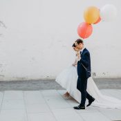 Hochzeitswahn - Eine Braut in einem fließenden weißen Kleid und ein Bräutigam in einem marineblauen Anzug gehen Hand in Hand an einer weißen Wand des Oberhauses entlang, der Bräutigam hält verspielt leuchtende Luftballons (rot Oberhaus