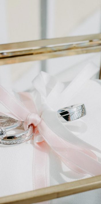 Hochzeitswahn - Ein Paar Eheringe, mit einer rosa Schleife zusammengebunden, liegt auf einem weißen Kissen auf einem Stuhl mit Goldrahmen, beleuchtet vom natürlichen Sonnenlicht während einer Traumhochzeit. Oberhaus