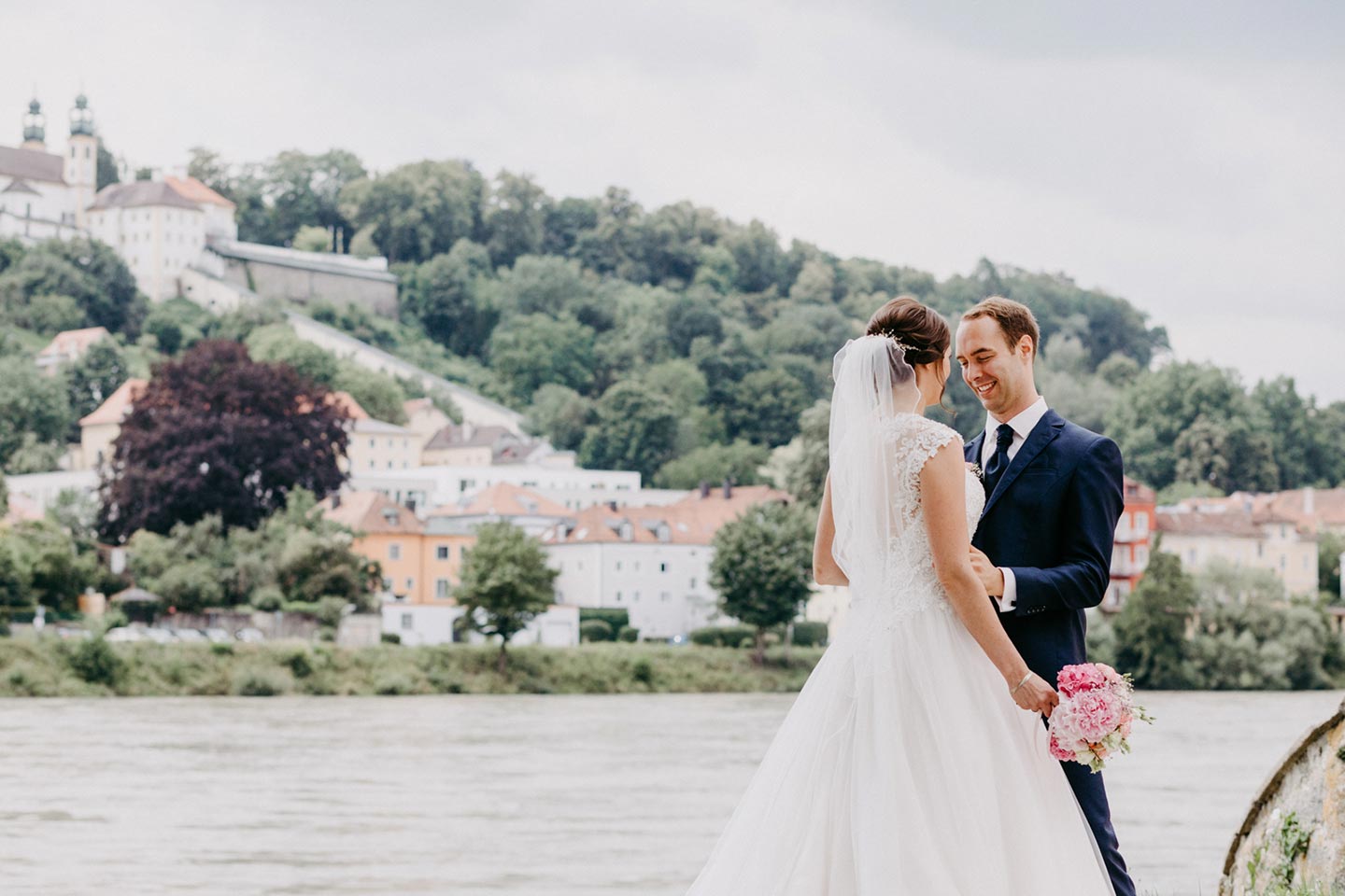 Hochzeitswahn - Braut und Bräutigam verbringen einen zärtlichen Moment am Flussufer, im Hintergrund die historischen Oberhausgebäude. Sie hält einen Blumenstrauß in der Hand; beide lächeln sanft. Oberhaus