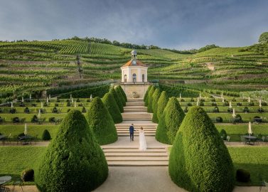 Hochzeitswahn - Ein Paar steht Hand in Hand in der Mitte eines symmetrischen Barockgartens, der unter klarem Himmel zu einem reich verzierten Pavillon inmitten üppiger, terrassierter Weinberge führt.