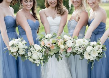 Hochzeitswahn - Eine Braut in Weiß und ihre Brautjungfern in passenden hellblauen Kleidern präsentieren ihre eleganten Blumensträuße, die mit einer zarten Mischung aus Rosen und Grünpflanzen gefüllt sind und den freudigen Geist der Hochzeitsfeier verkörpern.