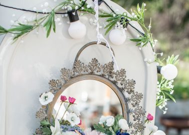 Hochzeitswahn - Ein eleganter Vintage-Spiegel, geschmückt mit einem farbenfrohen Blumenarrangement und Lichterketten, befestigt an einem klassischen weißen Fahrzeug, wahrscheinlich Teil einer festlichen Veranstaltung oder Feier im Freien.