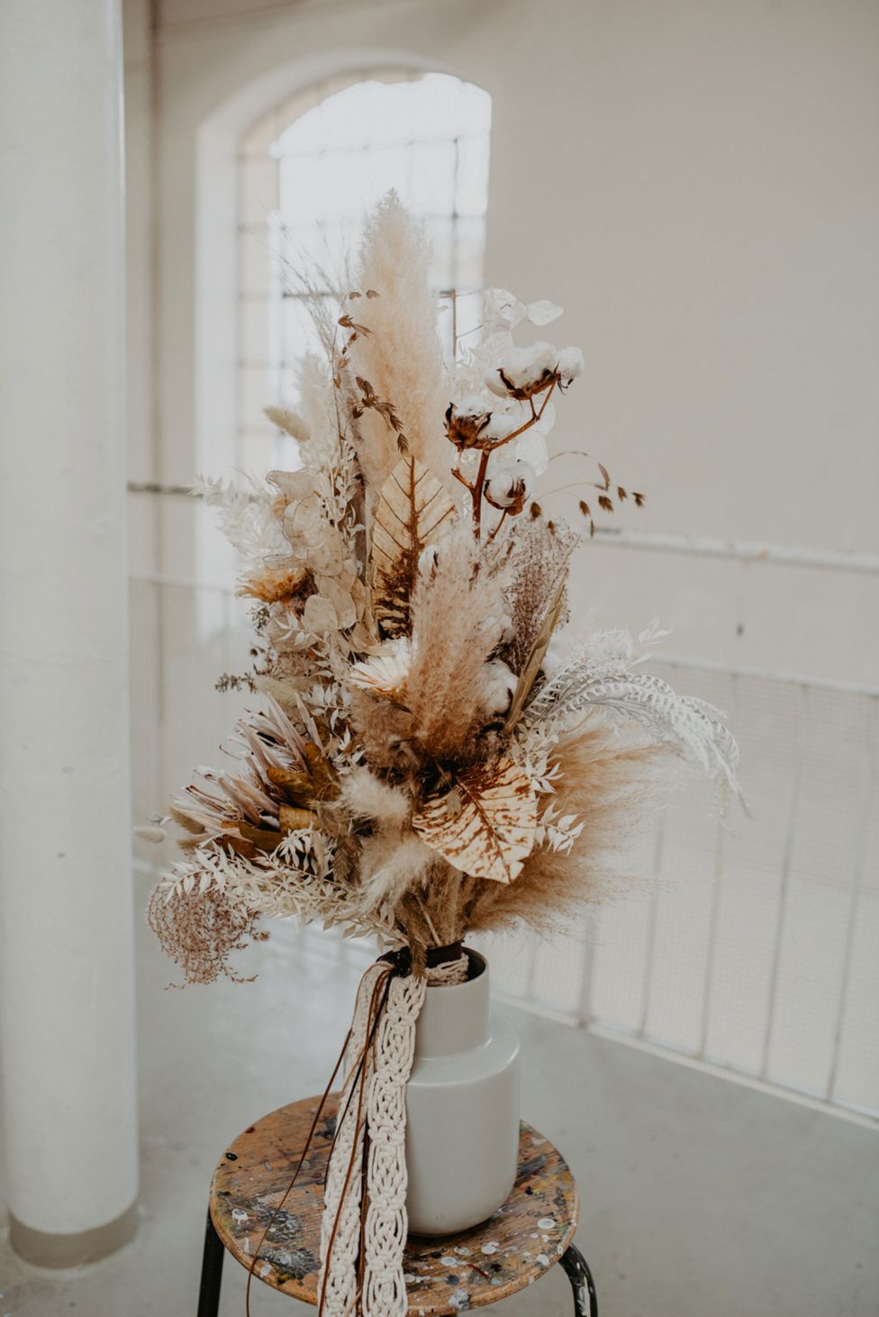 Hochzeitswahn - Ein Strauß getrockneter Pampasgras, Farne und Baumwollpflanzen in einer schlichten weißen Vase, auf einem alten Hocker vor einer cremefarbenen Wand platziert, strahlt rustikale Eleganz aus.