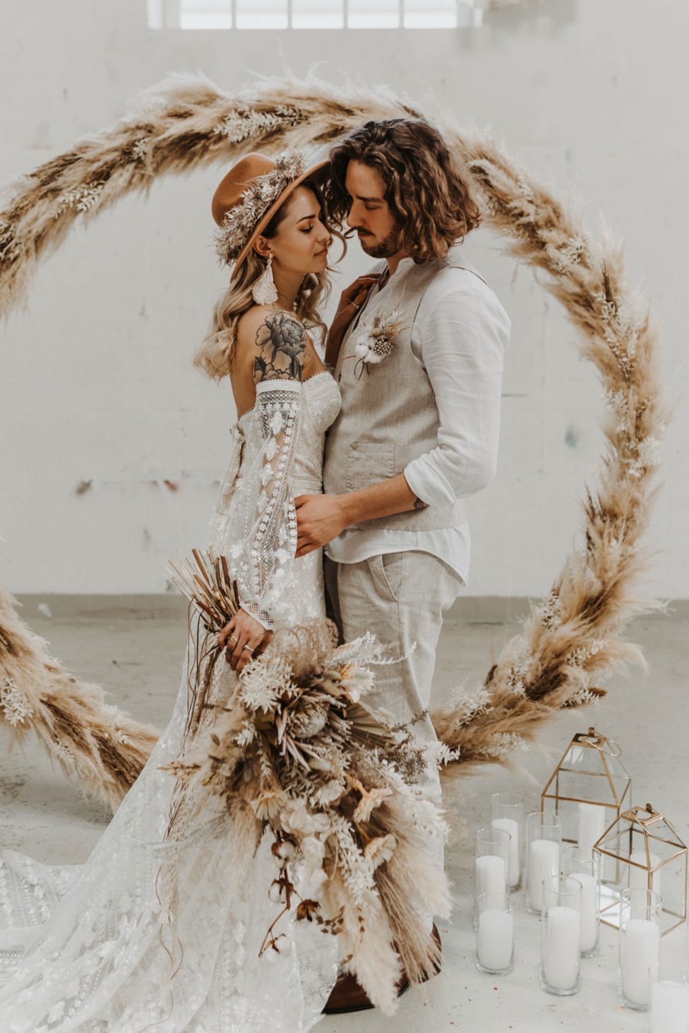 Hochzeitswahn - Ein Paar umarmt sich zärtlich in einer rustikalen Hochzeitskulisse, umgeben von einem Halbkreis aus Pampasgras, mit Kerzen zu ihren Füßen, und vermittelt so unkonventionelle Eleganz und intime Romantik.