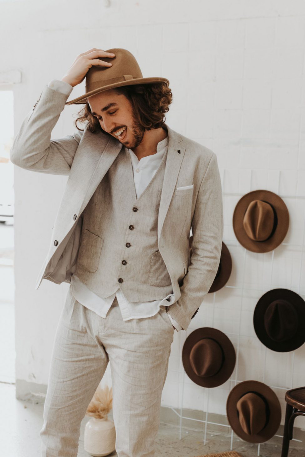 Hochzeitswahn - Ein eleganter Mann in grauem Anzug und weißem Hemd steht lächelnd und an seinen Hut lüpfend in einem Raum, an dessen Wand eine Reihe von Hüten hängt.