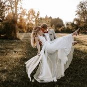 Hochzeitswahn - Ein Bräutigam im beigen Anzug hebt eine Braut im weißen Brautkleid hoch, wobei sie während ihrer böhmischen Hochzeit in einem sonnendurchfluteten, von Bäumen umgebenen Park verspielt den Fuß hochstreckt. Magical-Homes