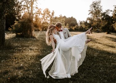 Hochzeitswahn - Ein Bräutigam im beigen Anzug hebt eine Braut im weißen Brautkleid hoch, wobei sie während ihrer böhmischen Hochzeit in einem sonnendurchfluteten, von Bäumen umgebenen Park verspielt den Fuß hochstreckt. Magical-Homes