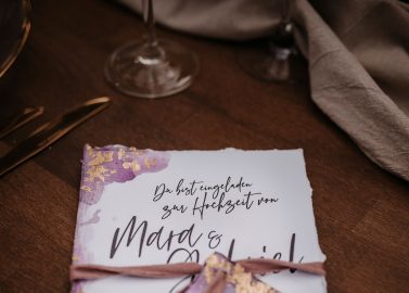 Hochzeitswahn - Auf einem Holztisch liegt eine Hochzeitseinladung mit eleganter violetter Schrift und Akzenten, zusammengebunden mit einem passenden Band. In der Nähe runden ein Glas Wein und eine Stoffserviette die herbstliche Verse ab.