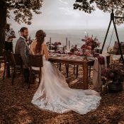Hochzeitswahn - Ein Hochzeitspaar sitzt an einem rustikalen, mit Blumenarrangements geschmückten Tisch im Freien an der Heuchelberger Warte mit Blick auf eine weite Landschaft. Die Braut in einem langen weißen Kleid und der Bräutigam sind