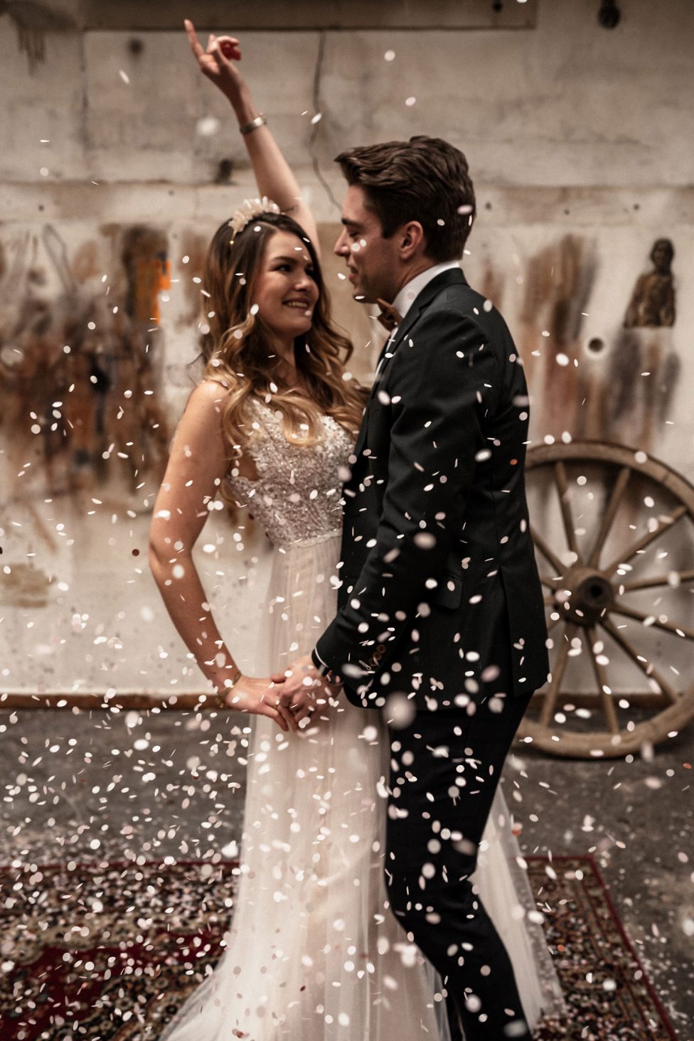 Hochzeitswahn - Ein fröhliches, frisch vermähltes Paar tanzt inmitten eines Konfettiregens, während die Braut begeistert den Arm streckt und der Bräutigam sie liebevoll ansieht. All dies fängt die festliche Atmosphäre ihrer Hochzeitsfeier ein.