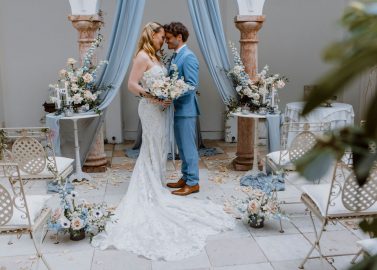 Hochzeitswahn - Ein glückliches Paar teilt einen zärtlichen Moment inmitten eines eleganten Dekors: Die Braut trägt ein elegantes weißes Kleid und der Bräutigam einen blauen Anzug, umgeben von ruhigen Blumen und klassischen Säulen, und feiert seinen Hochzeitstag.